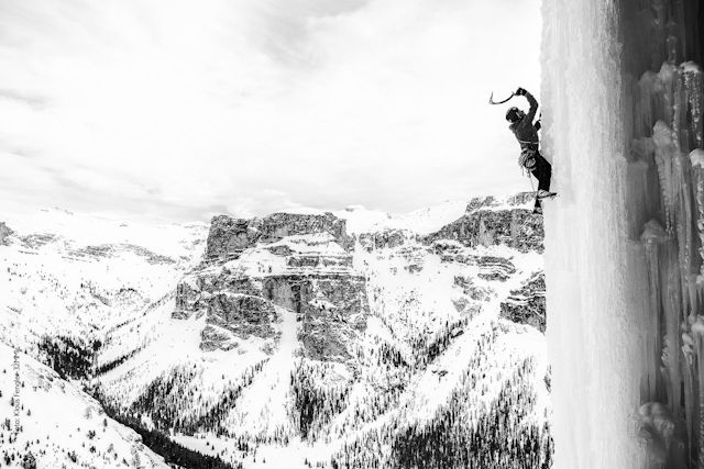En un primer plano, sobre una pared de hielo vemos a un escalador usando un piolet para poder subir. Al fondo un macizo montañoso cubierto de nieve