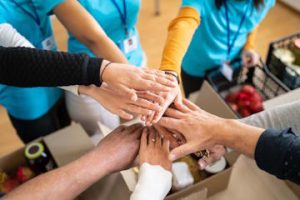 Miniatura, varias personas, de diferentes etnias edades, unen las manos sobre cajas con comida